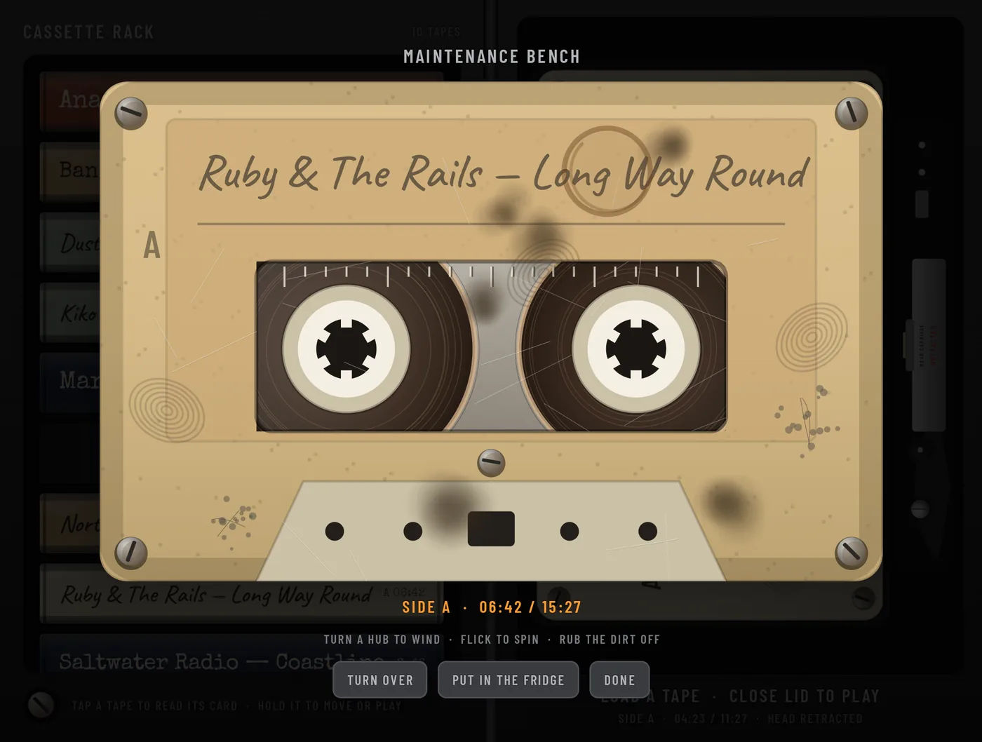 The maintenance bench: a large, yellowed and scratched cassette, Ruby and The Rails, Long Way Round, with dirt smudges, side A 06:42 of 15:27, the hint turn a hub to wind, flick to spin, rub the dirt off, and the keys TURN OVER, PUT IN THE FRIDGE and DONE.